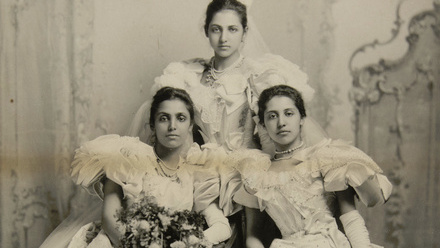 Posed group photograph of three young Asian women in grand white Victorian dresses.