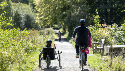 Two people cycling down a trail bordered by trees and plants. One on a recumbent tricycle.
