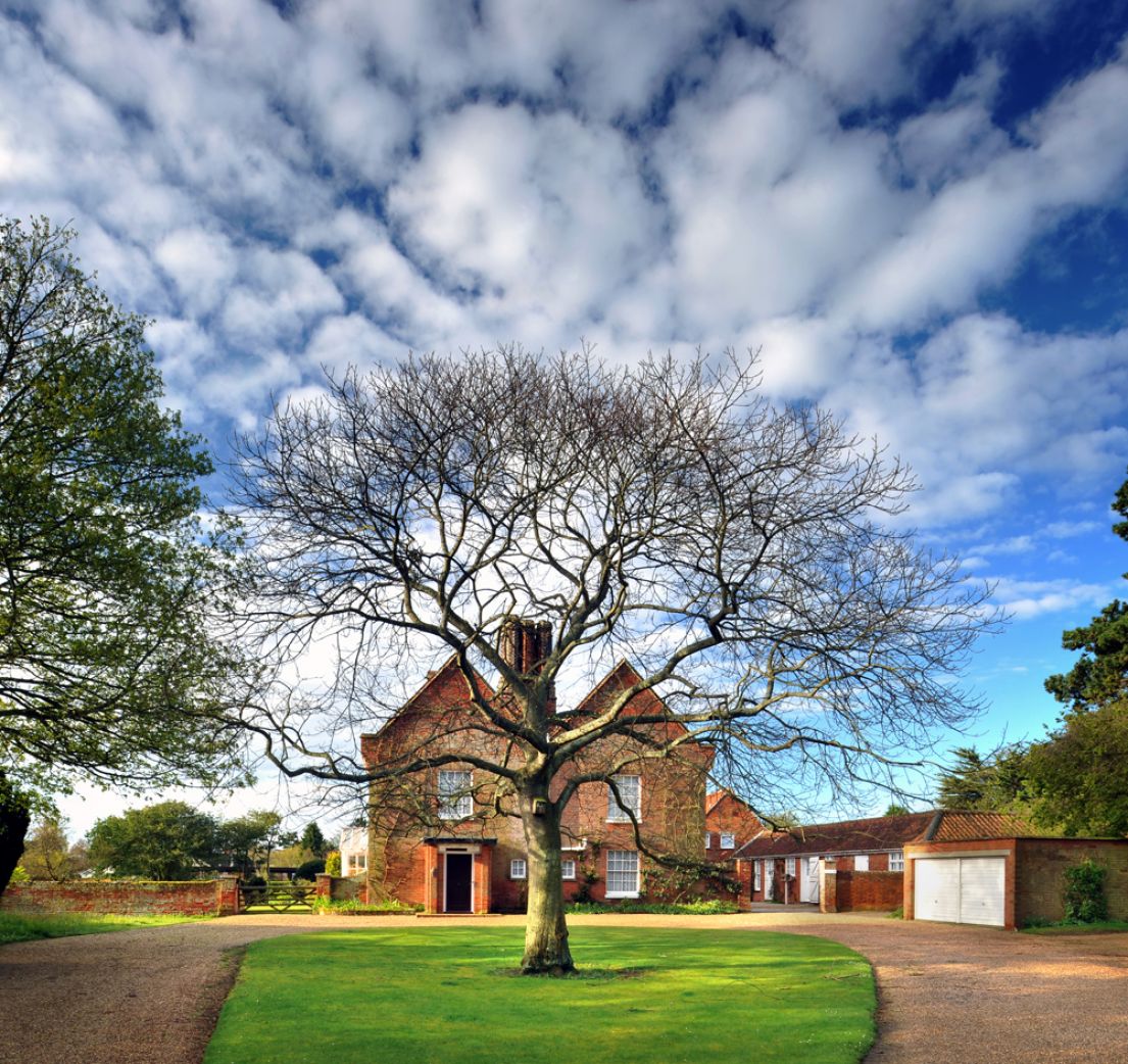 Looking up from the drive, with a circle of grass with a bare tree, up towards a large 17th-century red brick farmhouse.
