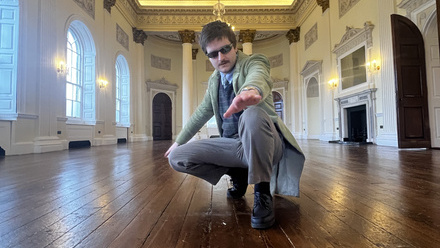 Man in pale suit and sunglasses crouched on wooden floor of a large, empty, sunlit, historic room with arched decorative ceiling.