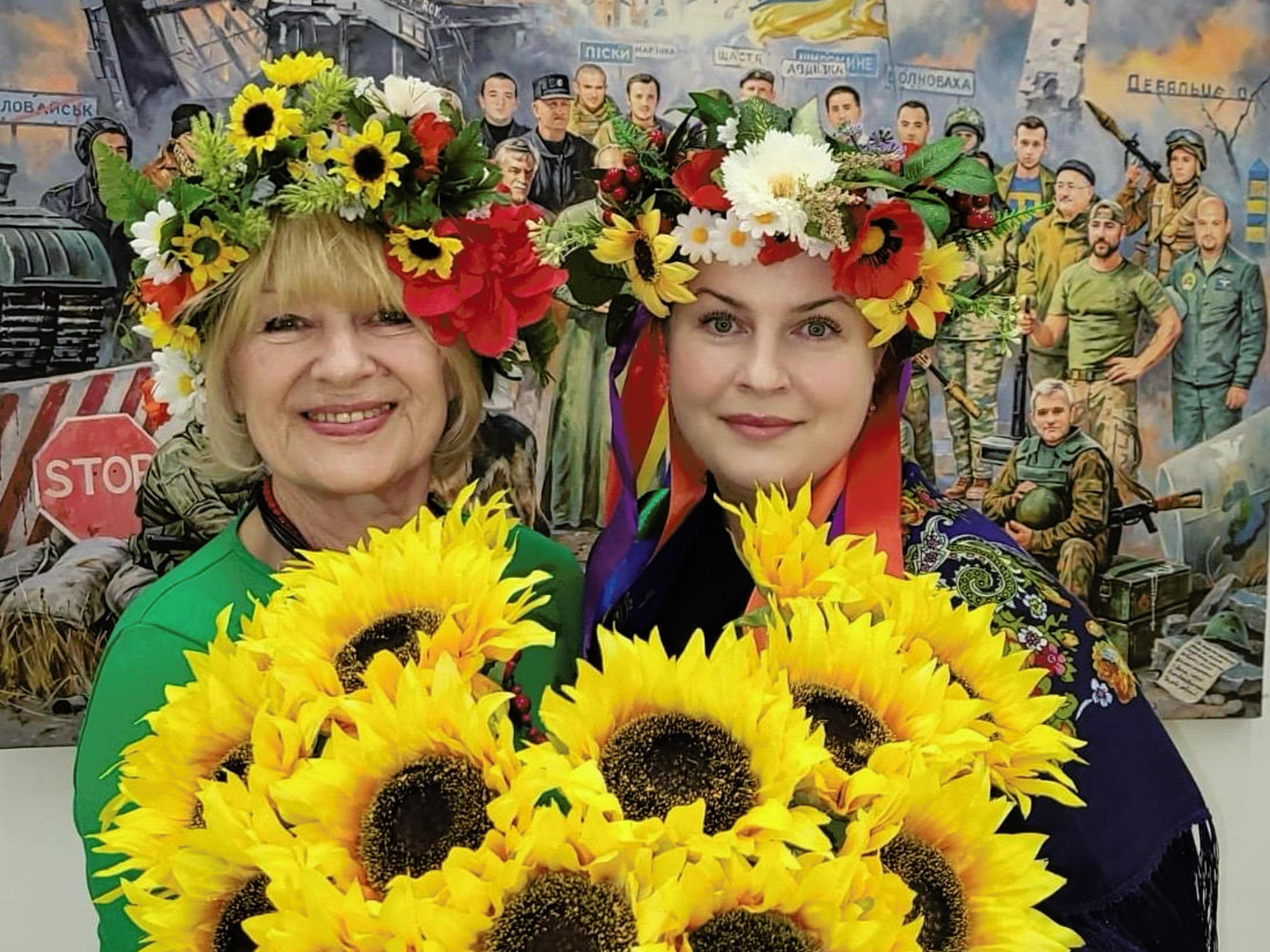 Two women in floral garland crowns holding a large bunch of sunflowers.