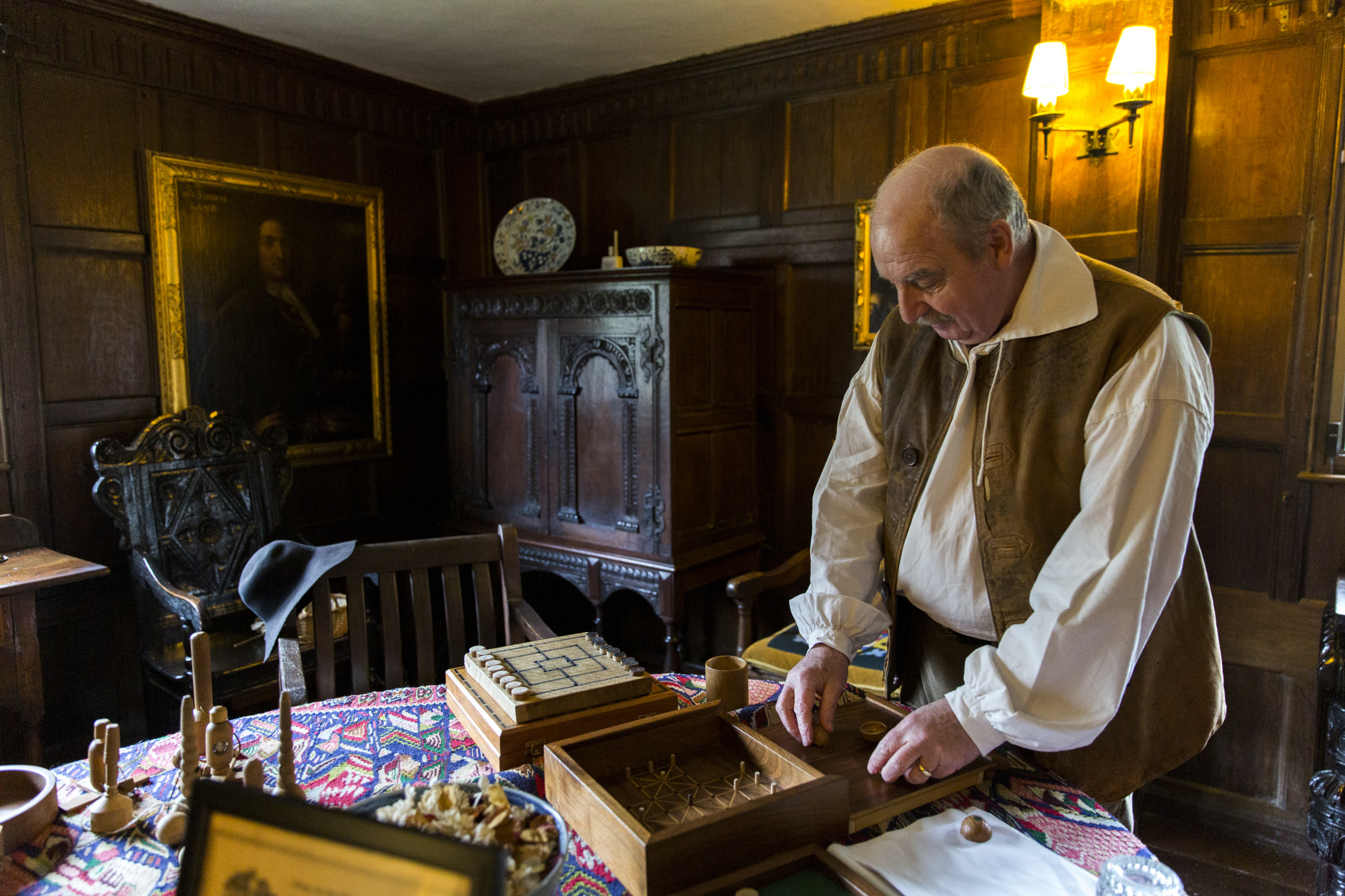 Man in medieval costume sorting through a wooden gaming box on a table in a wood panelled room.