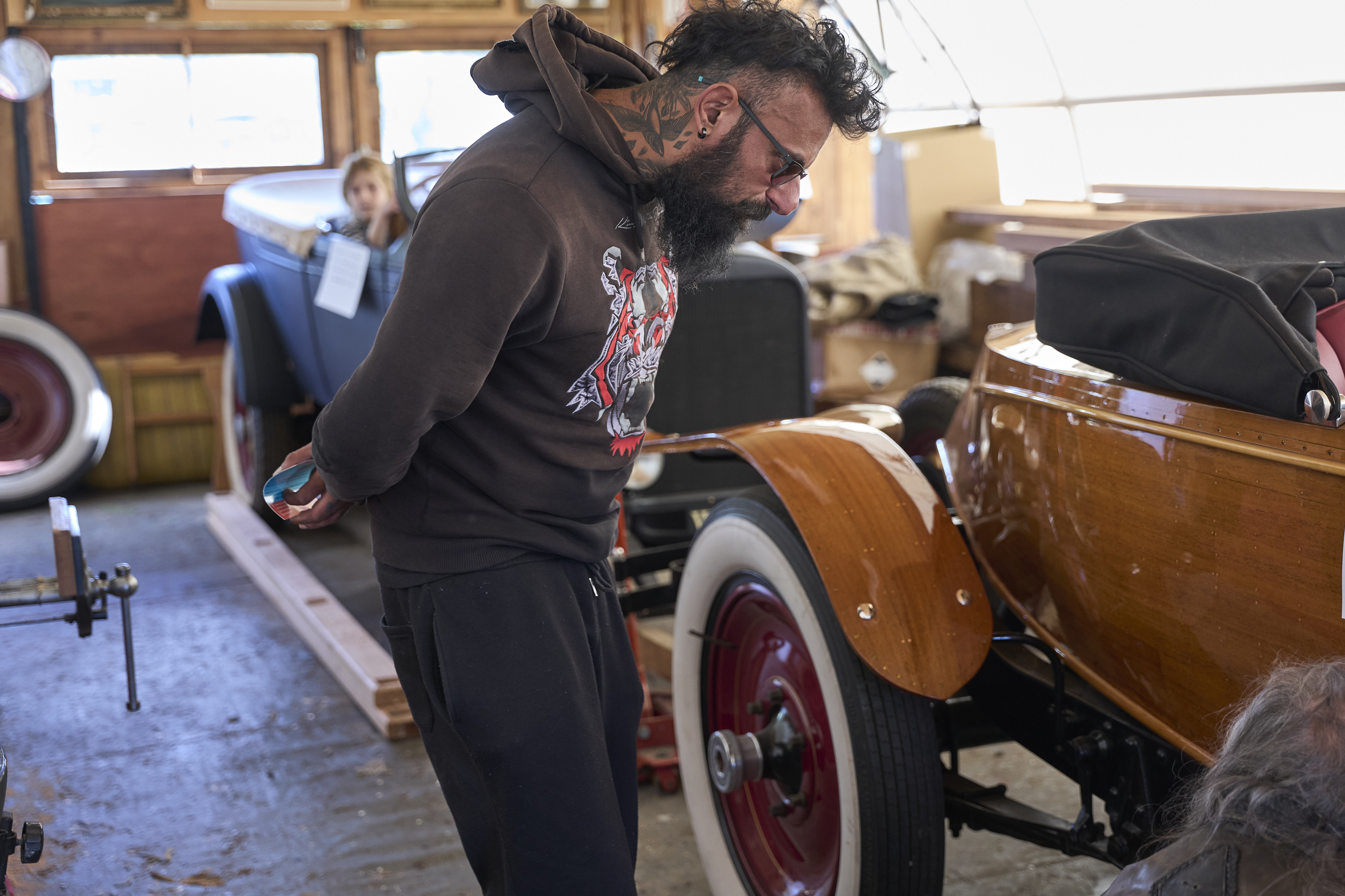 Bearded man in a hoodie looking at a vintage soft top car in a barn.