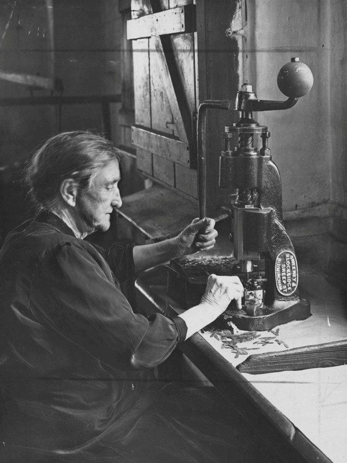 Older woman with hair in a bun sat working a metal punch machine at a table.