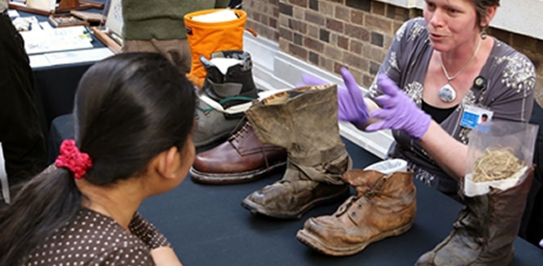 A woman with short red hair wearing purple latex gloves discussing old boots on the table to another woman with her hair tied up in a red scrunchie.