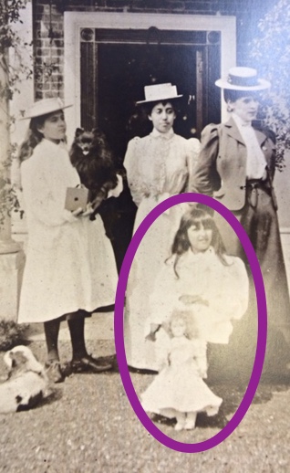 Victorian photograph of a woman stood outside a house with three girls of varying ages, the youngest holding a doll is circled in red.