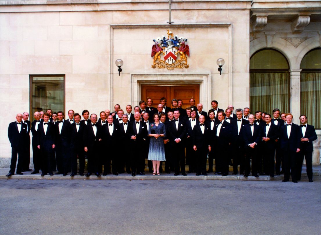 A staff photograph taken outside of a building, with men in suites and the sole female partner at Herbert Smith Freehills stood at the front in blue.