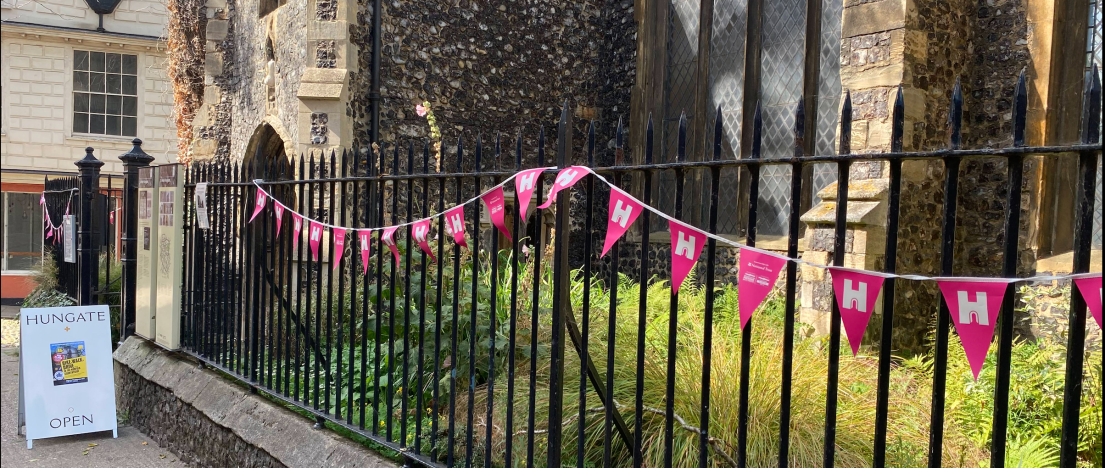 An old stone church. There is grass in front, with black metal railings adorned with pink and white Heritage Open Days bunting.