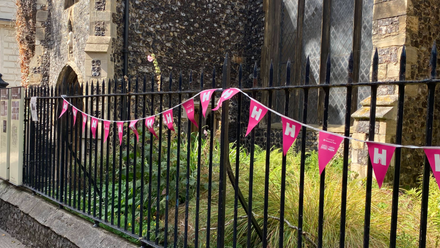 An old stone church. There is grass in front, with black metal railings adorned with pink and white Heritage Open Days bunting.