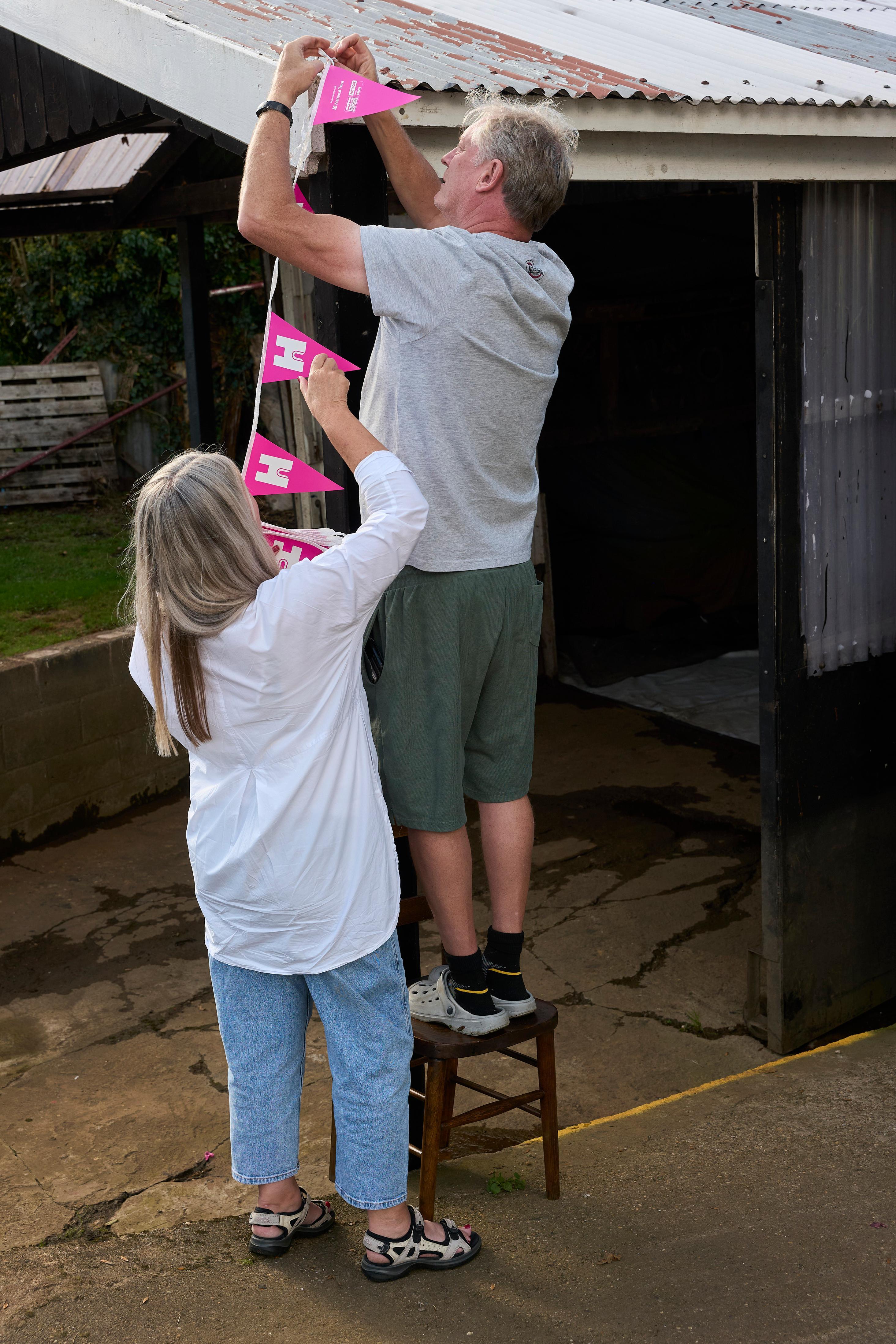 Man stood on chair pinning bunting to a corrugated roof, assisted by a woman.