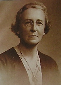 Sepia photographic portrait of older woman wearing glasses in a dark cardigan (or dress) with a pendant necklace.