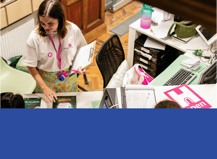 Person in white t-shirt, with pink lanyard holding a clipboard standing behind a desk speaking to visitors. 