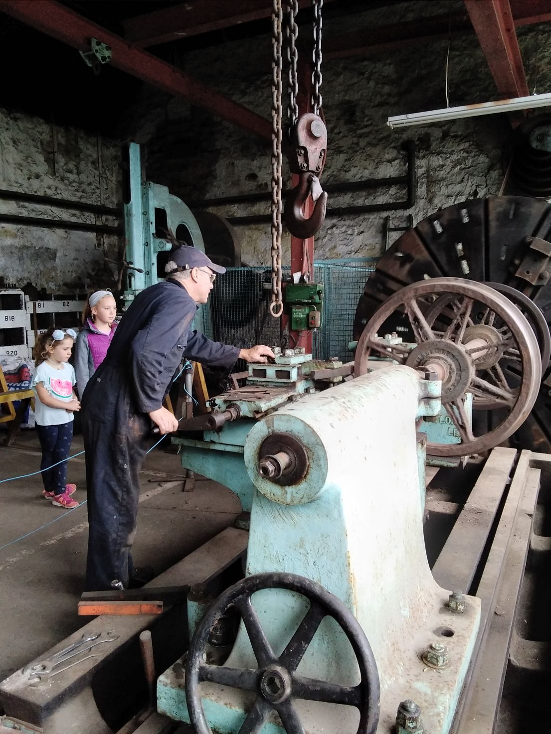 A man in dirty navy blue work overalls and cap demonstrating machinery with wheels, to two girls who look on.
