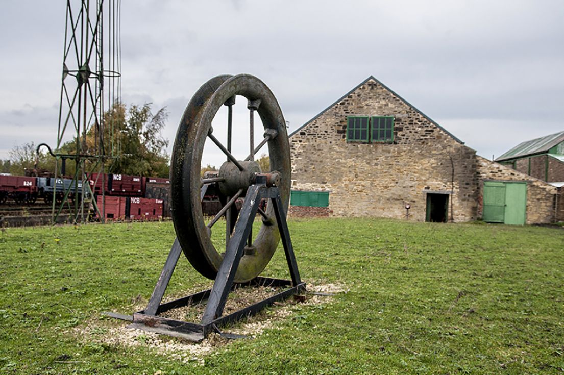 A stationary mounted Pit Wheel - a wheel sitting in an axis, to the left railway tracks and carriages, to the right an old stone building.