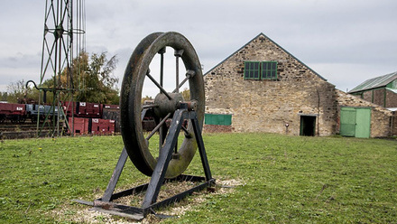 A stationary mounted Pit Wheel - a wheel sitting in an axis, to the left railway tracks and carriages, to the right an old stone building.