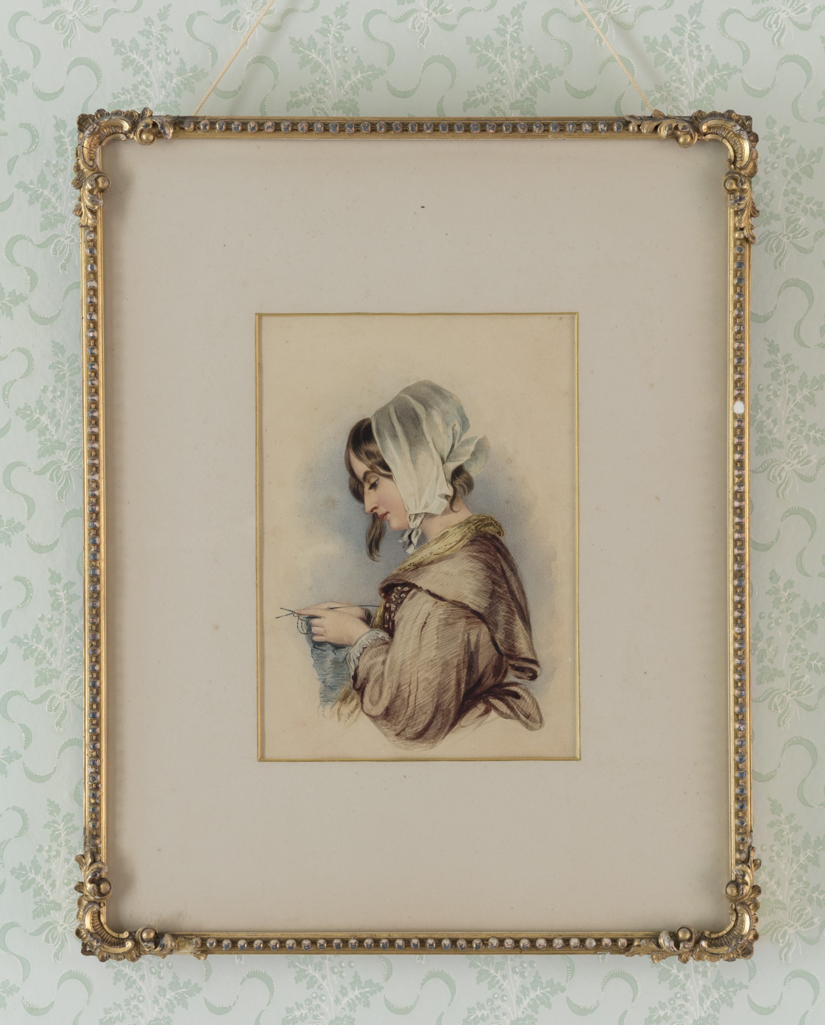 Watercolour portrait of a woman in a white cap knitting, hung on a patterned wall in a gold embossed frame.