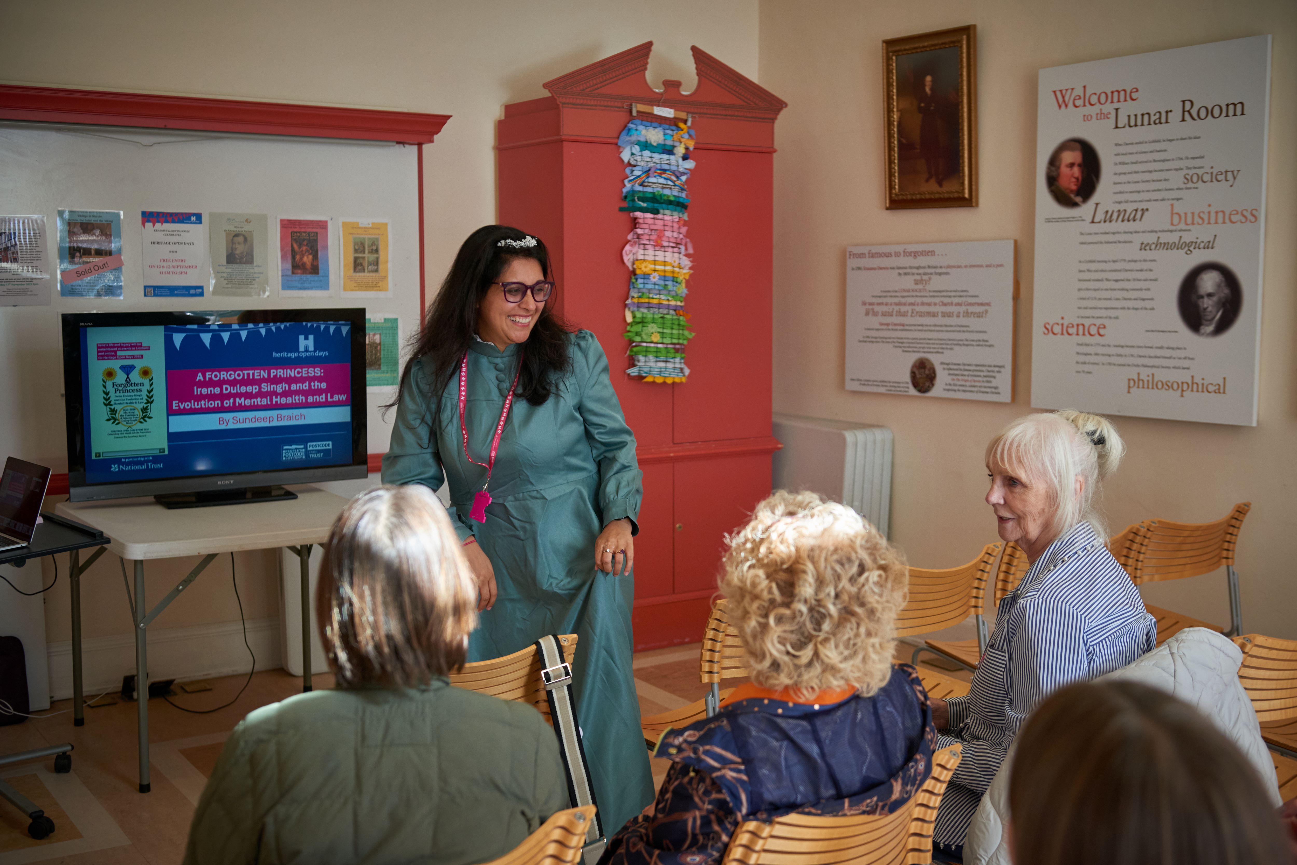 Young woman stood in front of a small tv screen talking with a group of older adults sat on chairs in a small room.