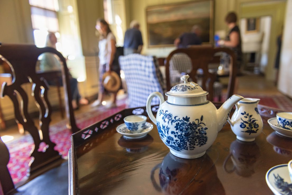 Focused white tea pot with intricate blue design set on a dark wooden table. Around it, matching jugs and cups on sauces with a similar blue pattern.