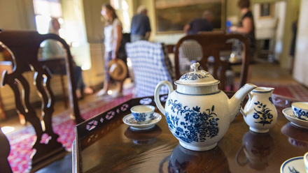 Focused white tea pot with intricate blue design set on a dark wooden table. Around it, matching jugs and cups on sauces with a similar blue pattern.