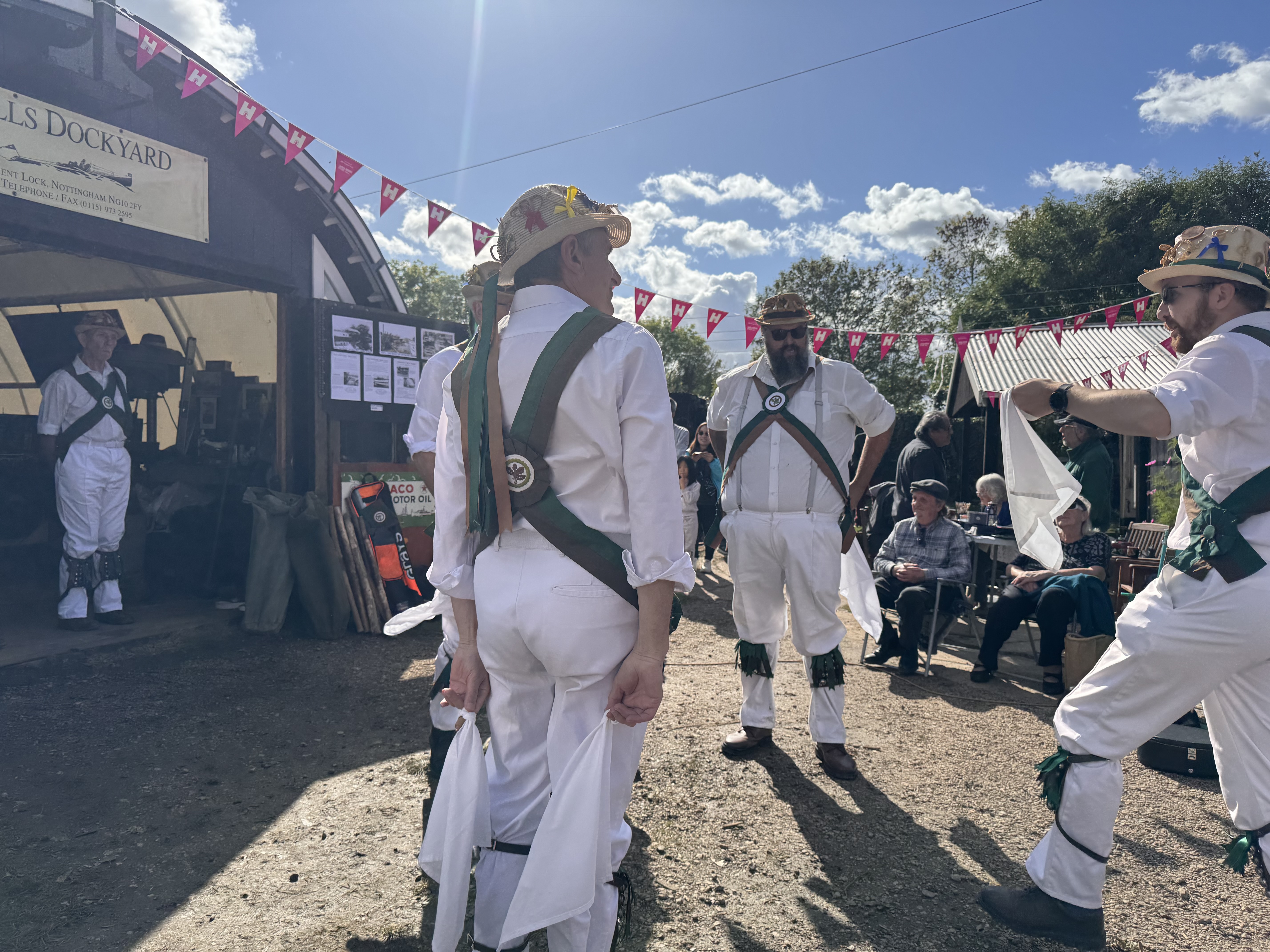 Morris dancers outside a large metal shelter with arched roof, pink bunting strung across in the background.
