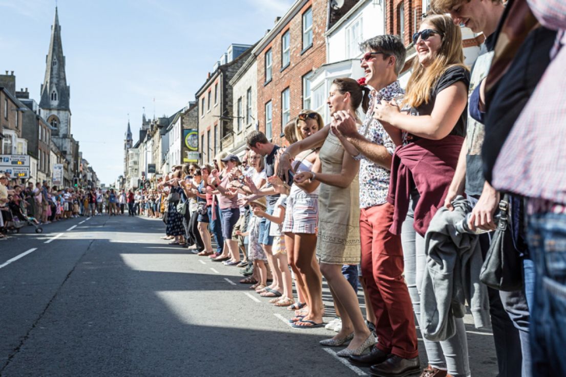 Many people lining a street in a town centre, dancing the Hokey Cokey.