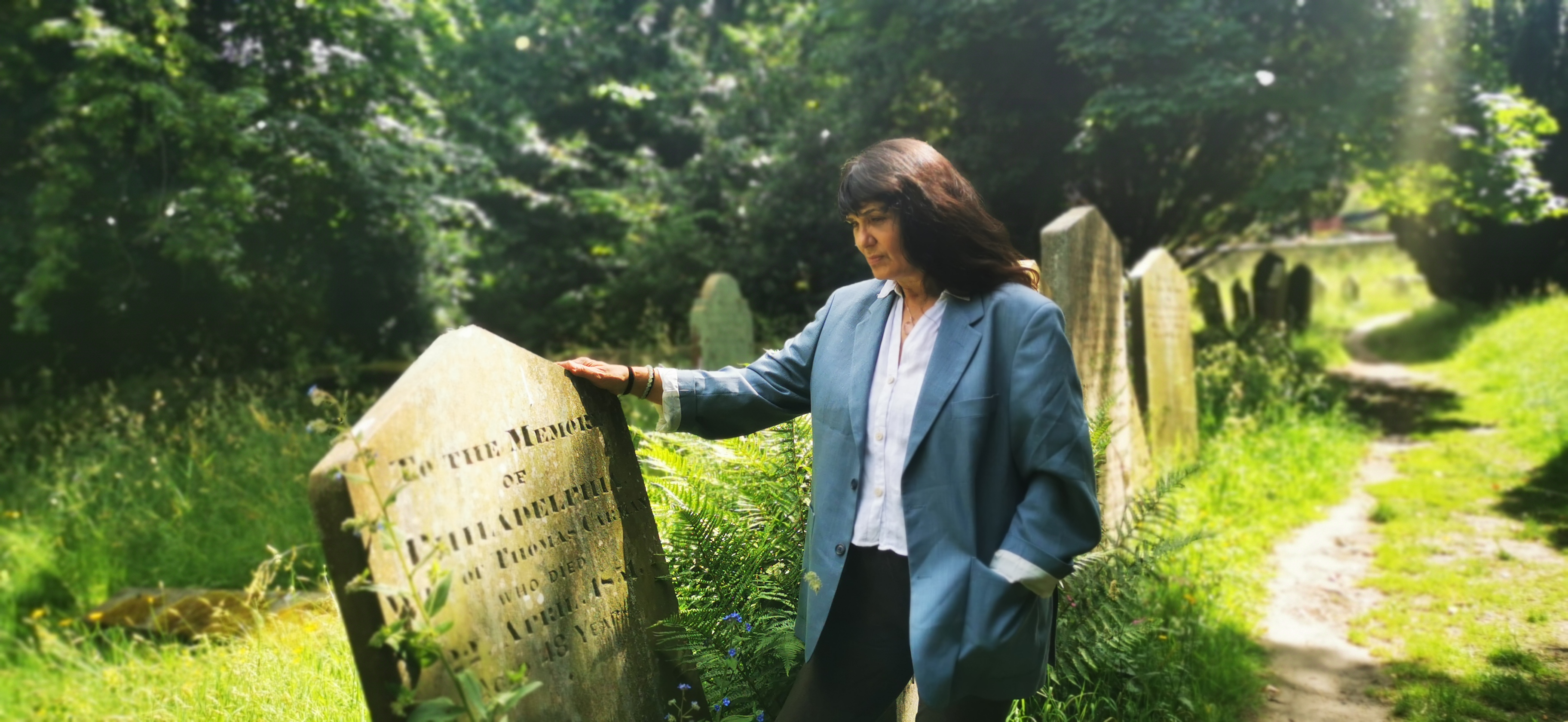 Woman in blue jacket stood beside a tilted headstone in a green cemetery park.