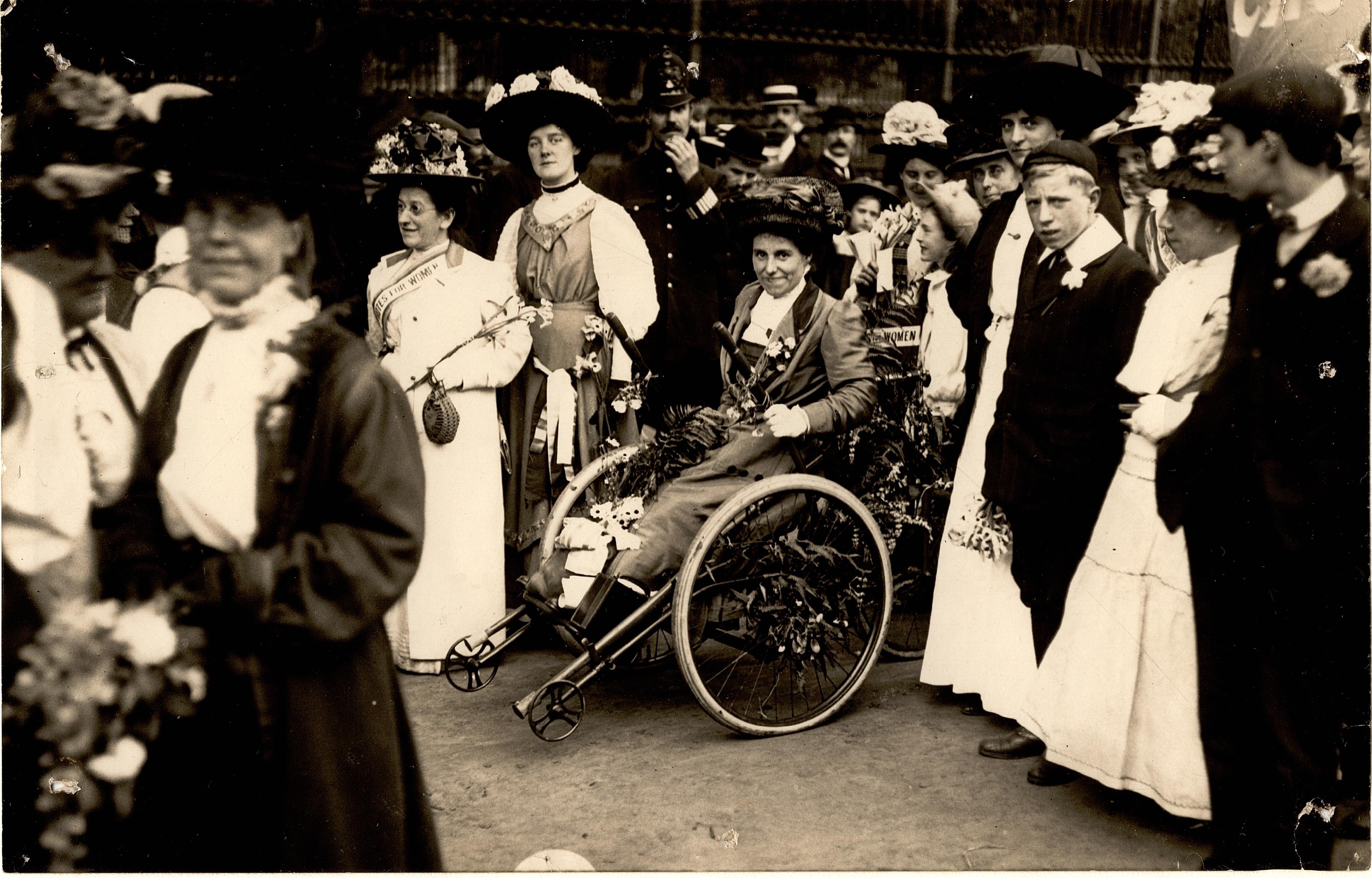 Smartly dressed Victorian woman with flowers in her lap, in a wheelchair amid a crowd.