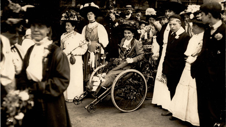 Smartly dressed Victorian woman with flowers in her lap, in a wheelchair amid a crowd.