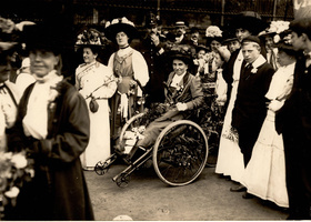 Smartly dressed Victorian woman with flowers in her lap, in a wheelchair amid a crowd.