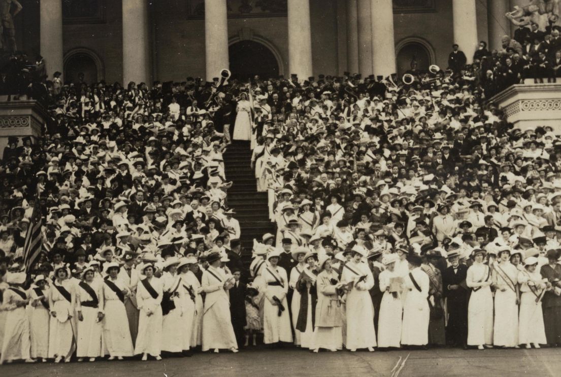 A black and white image of Suffragists wearing white dresses, hats and dark sashes, stood on the steps of the Capitol in Washington.