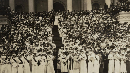 A black and white image of Suffragists wearing white dresses, hats and dark sashes, stood on the steps of the Capitol in Washington.