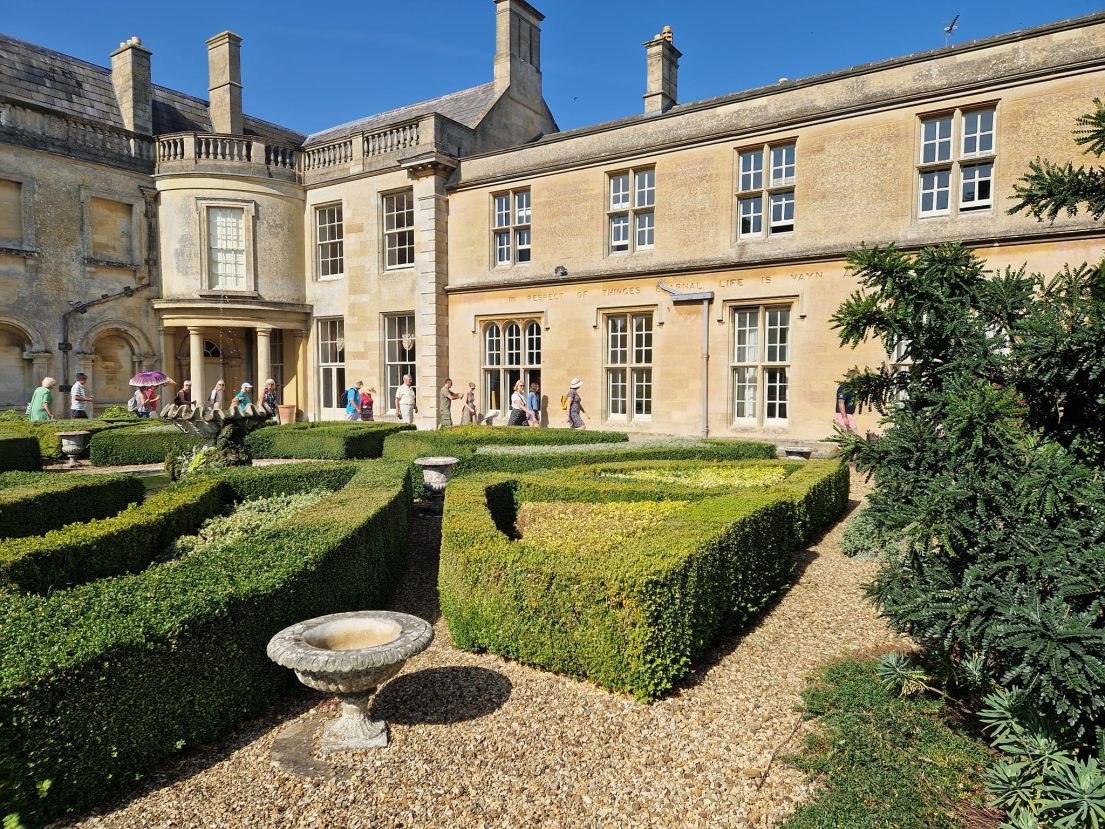 People walking in front of a two story stone building with a formal box hedge garden in the foreground.