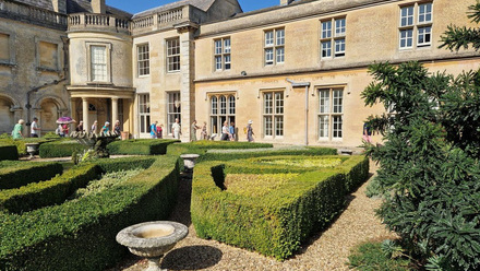 People walking in front of a two story stone building with a formal box hedge garden in the foreground.