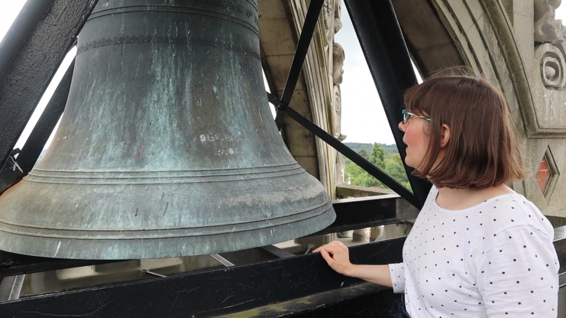 A women with short brown hair and white spotted top, examining a bell in an open belfry.