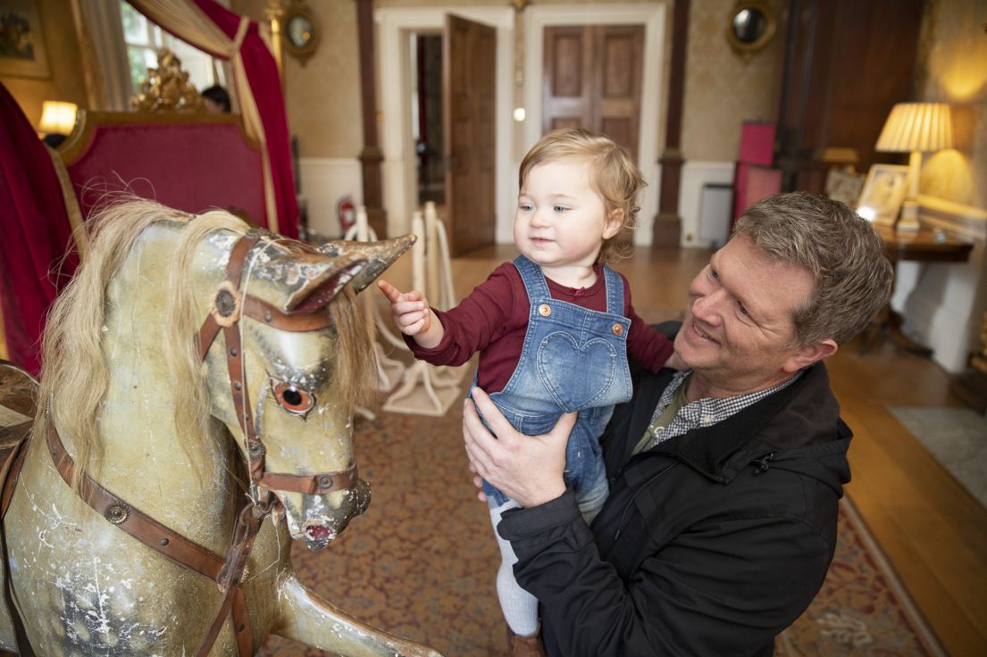 A small child being lifted up by an adult to examine a beautifully decorated wooden Victorian rocking horse. 