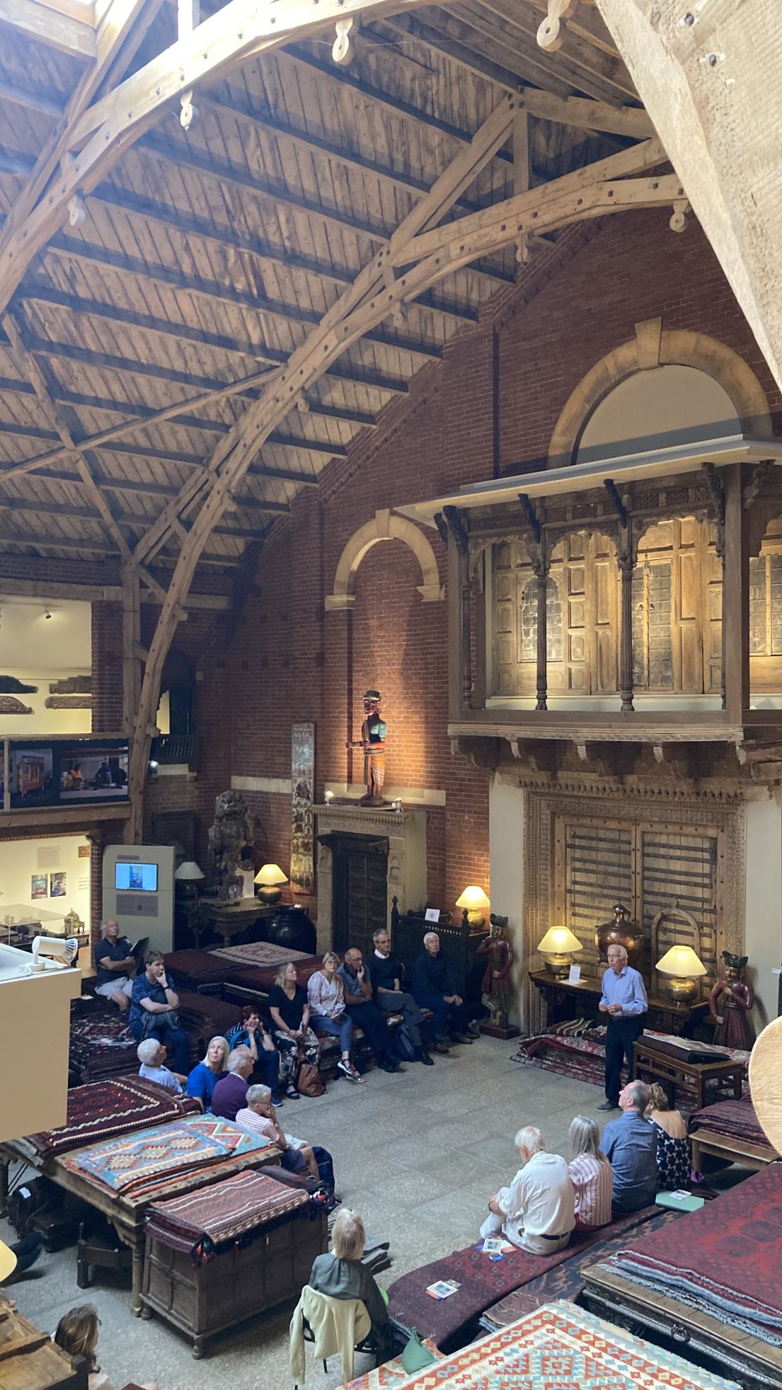 View from a gallery down into a cavernous hall with arched wooden roof. A group of people are sat in a square space below surrounded by artefacts.