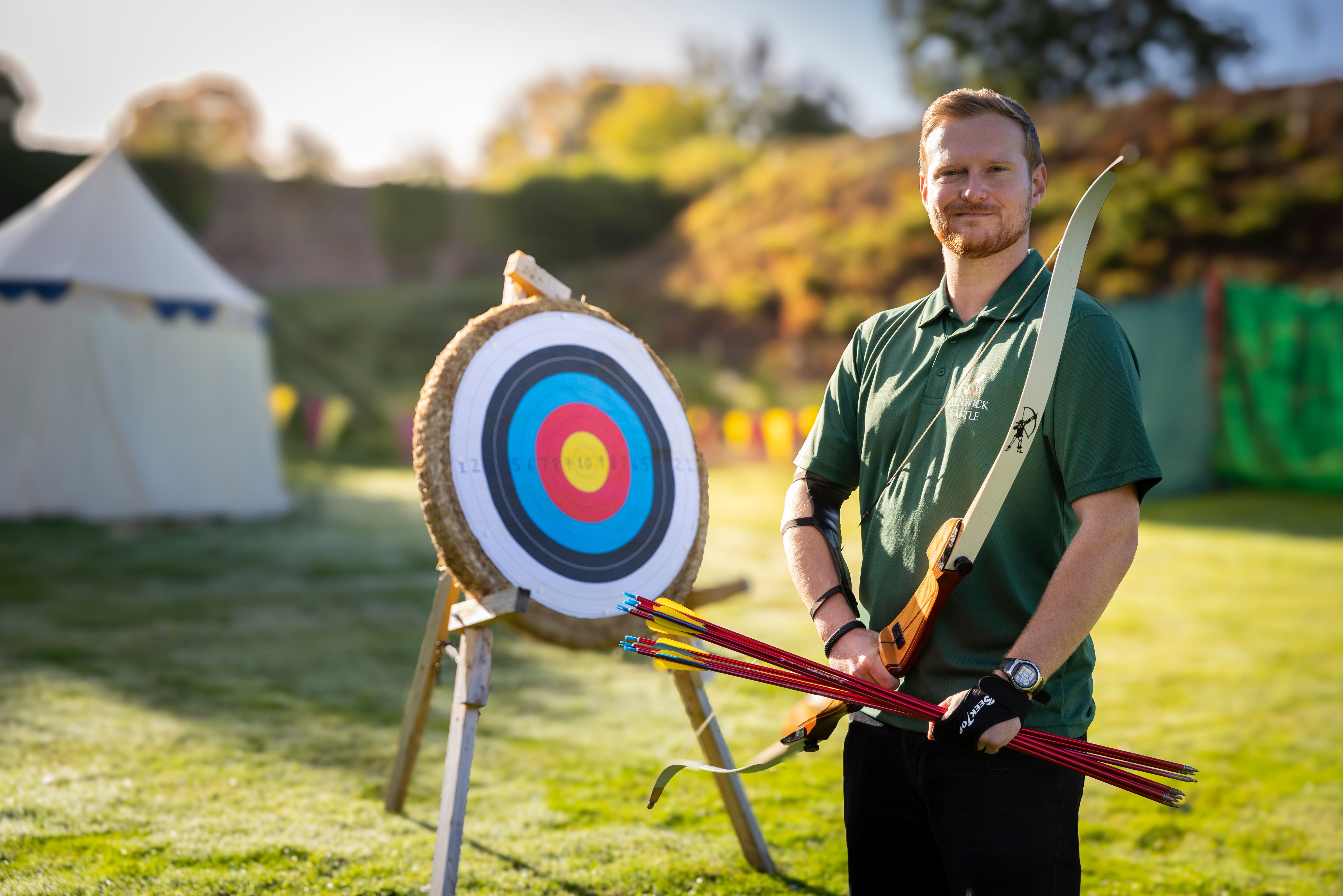 Man holding bow and arrows by a target board in a green space, with a tent in the background.