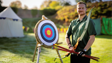 Man holding bow and arrows by a target board in a green space, with a tent in the background.