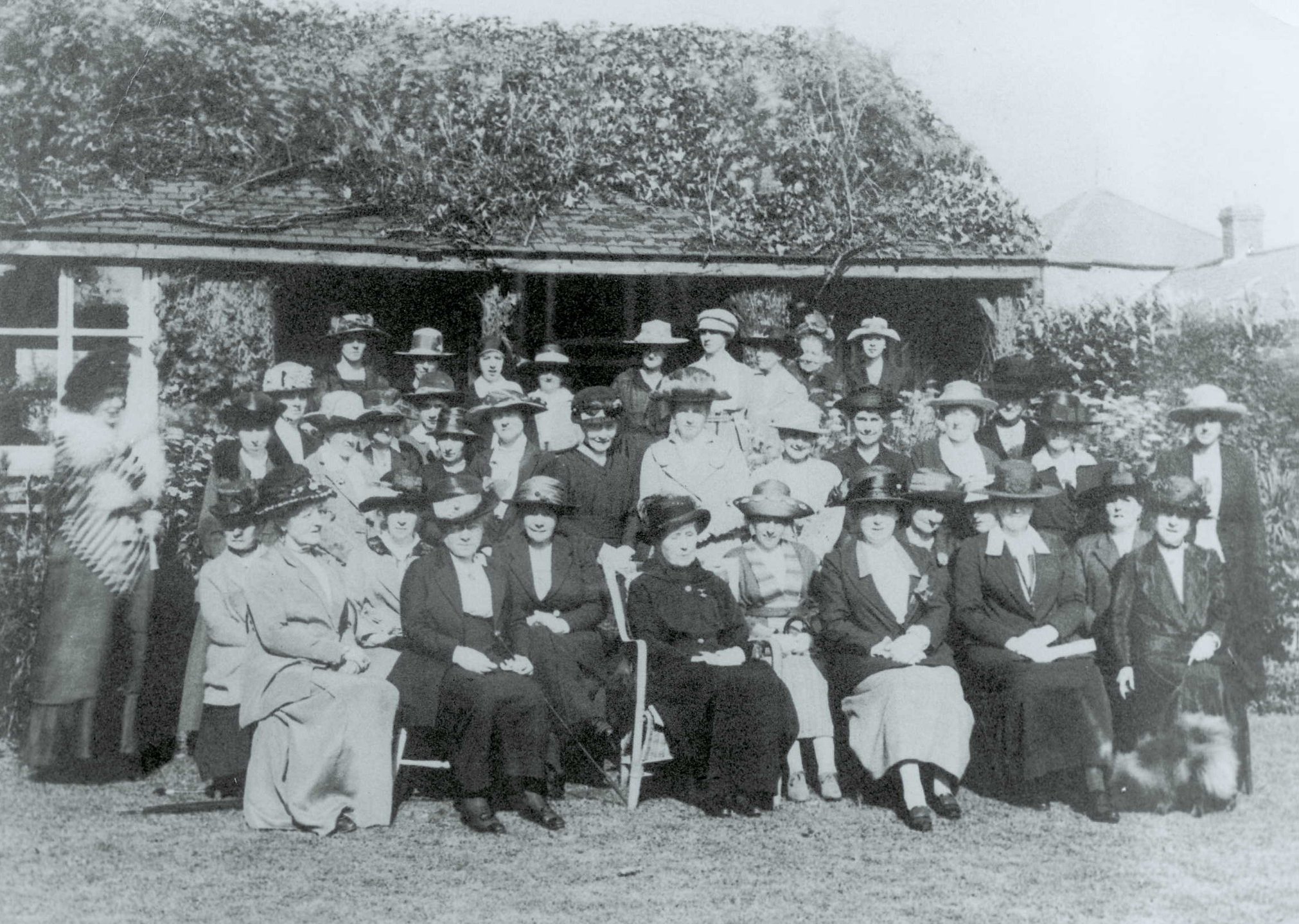 Old photo of a large group of women in hats seated/standing in a posed group in front of a low building with an overgrown tiled roof.