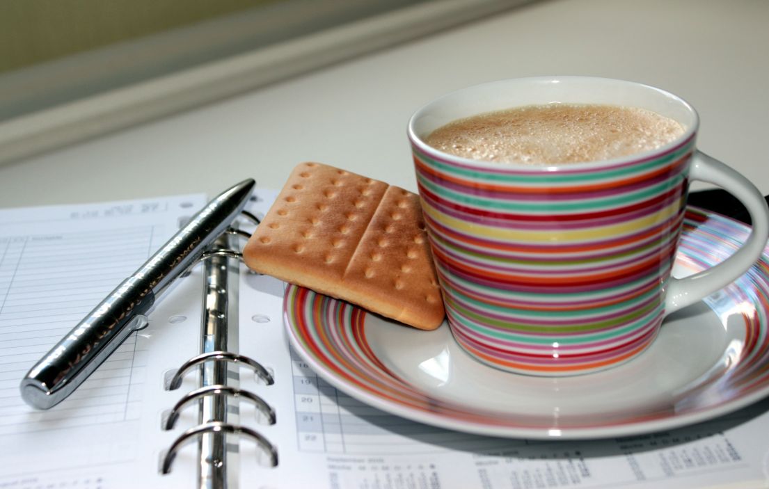 A cup of coffee and biscuit, placed on top of a diary calendar with a pen ready for notes. 