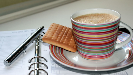 A cup of coffee and biscuit, placed on top of a diary calendar with a pen ready for notes. 