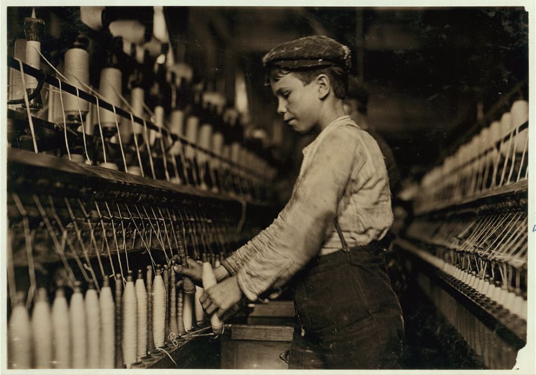 An old black and white photo of a young boy, wearing a cap working on and monitoring an industrial cotton machine.