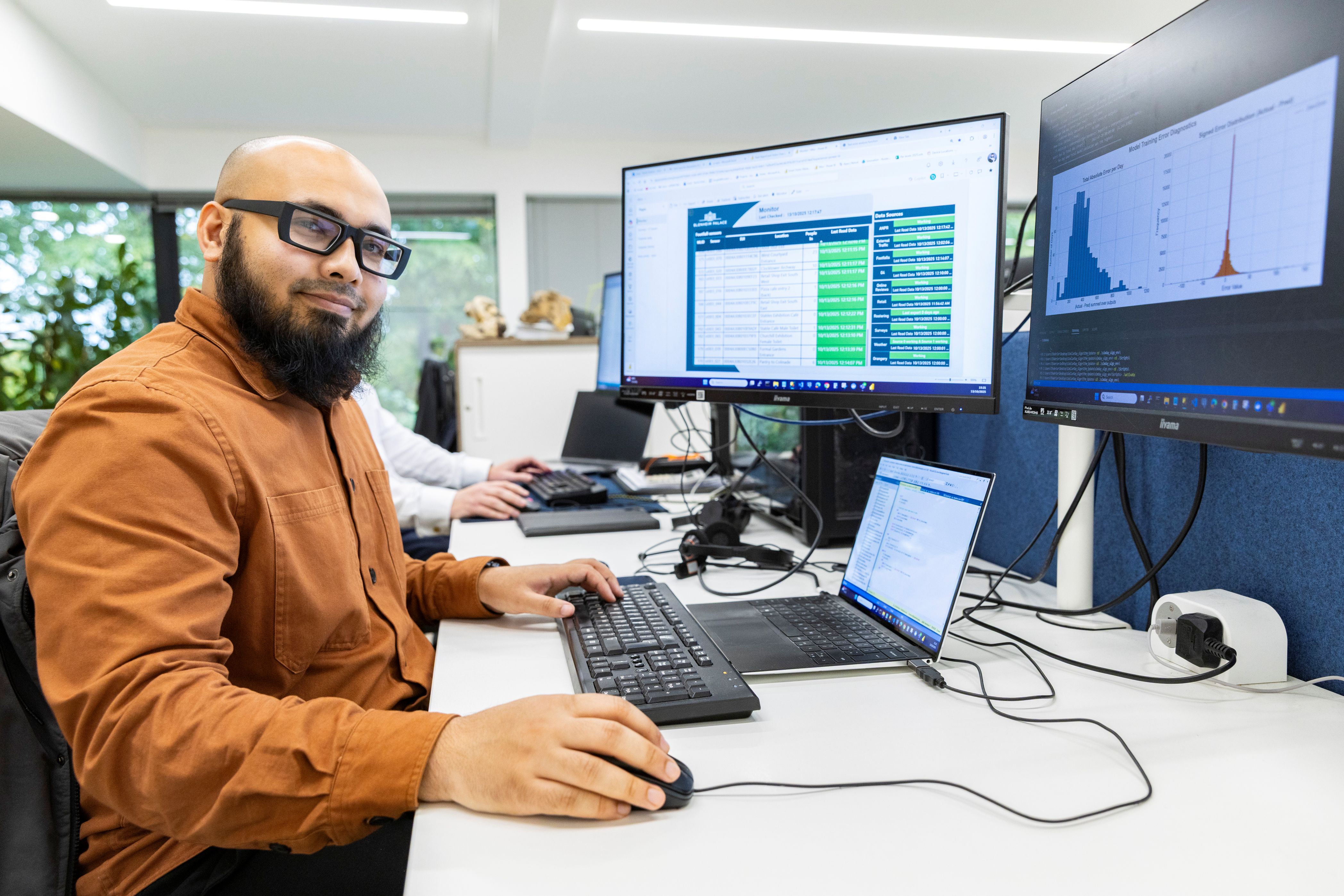 Man sat at modern office desk with laptop and 2 large monitors.
