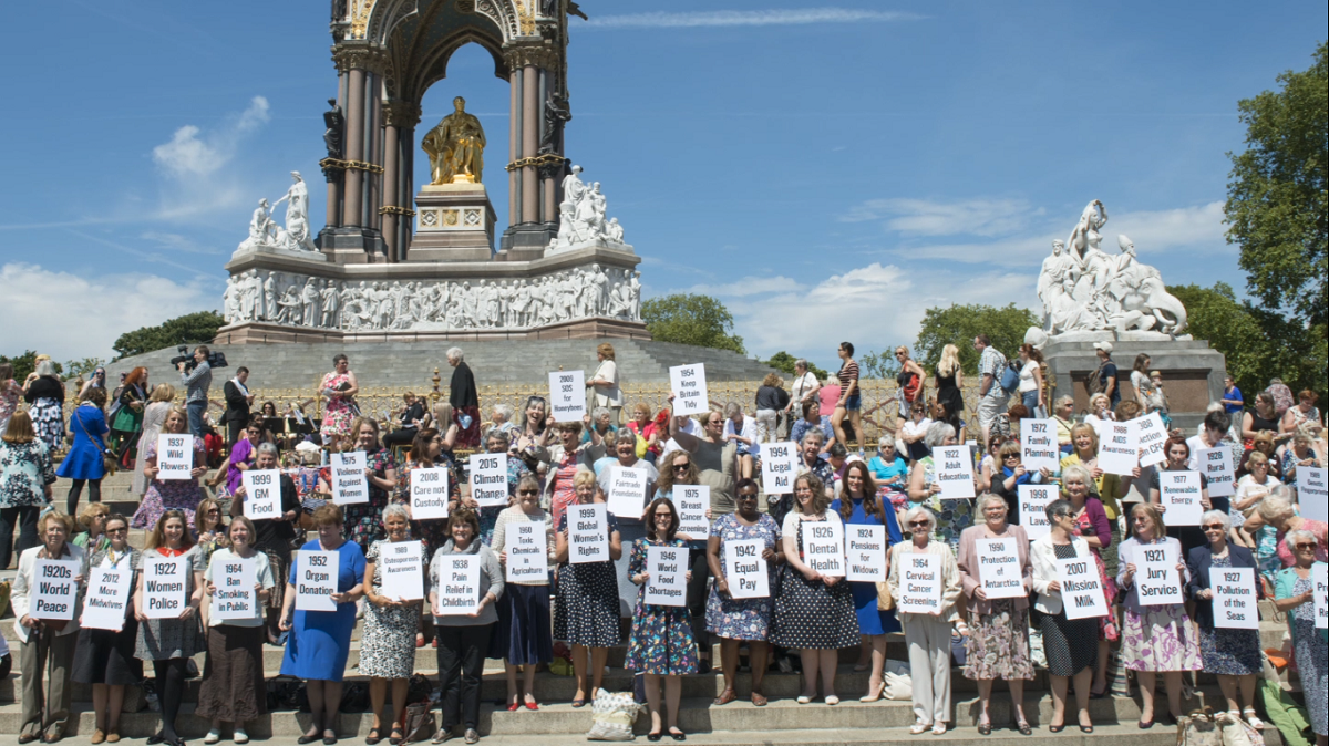 Large group of women holding white placards on the steps of a monument.