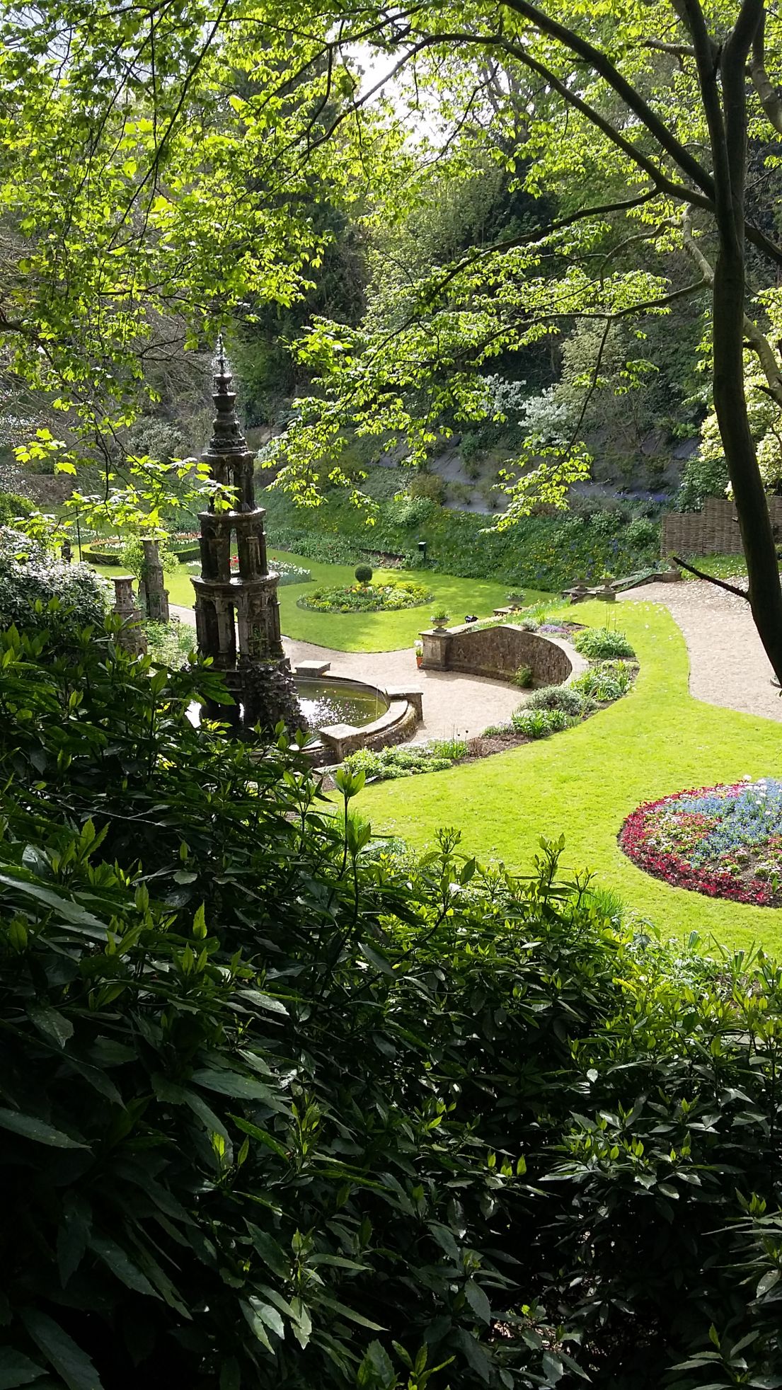 Looking down into a carefully designed garden with a tall fountain, circular flower beds and gravel paths.