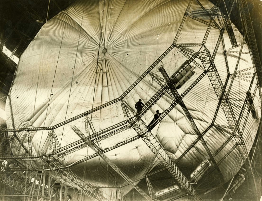 A black and white image of an airship, surrounded by scaffolding with workers helping with it's construction.