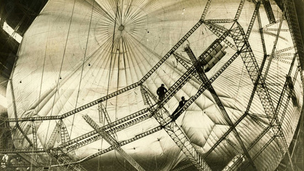 A black and white image of an airship, surrounded by scaffolding with workers helping with it's construction.