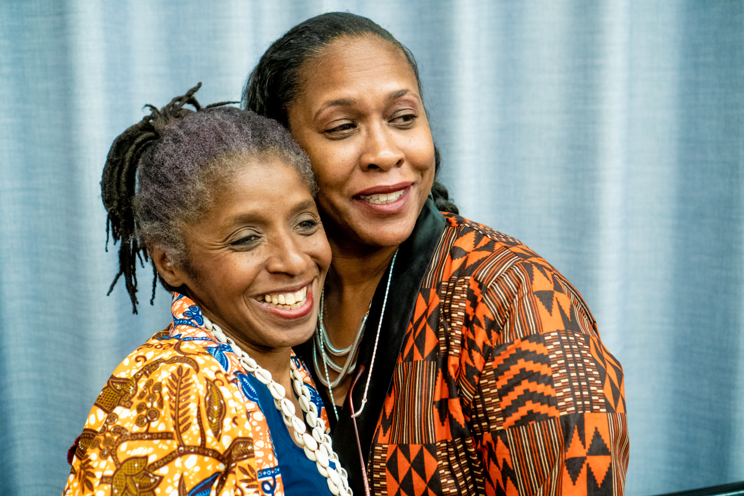 Two Black women in colourful outfits posing together in front of a blue curtain.