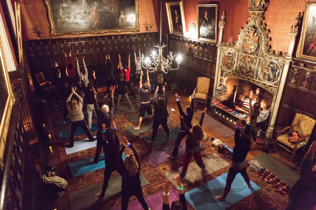An image taken from a height, looking down into a wooden panelled elaborate decorated room, with large portraits hung up, with people doing yoga.