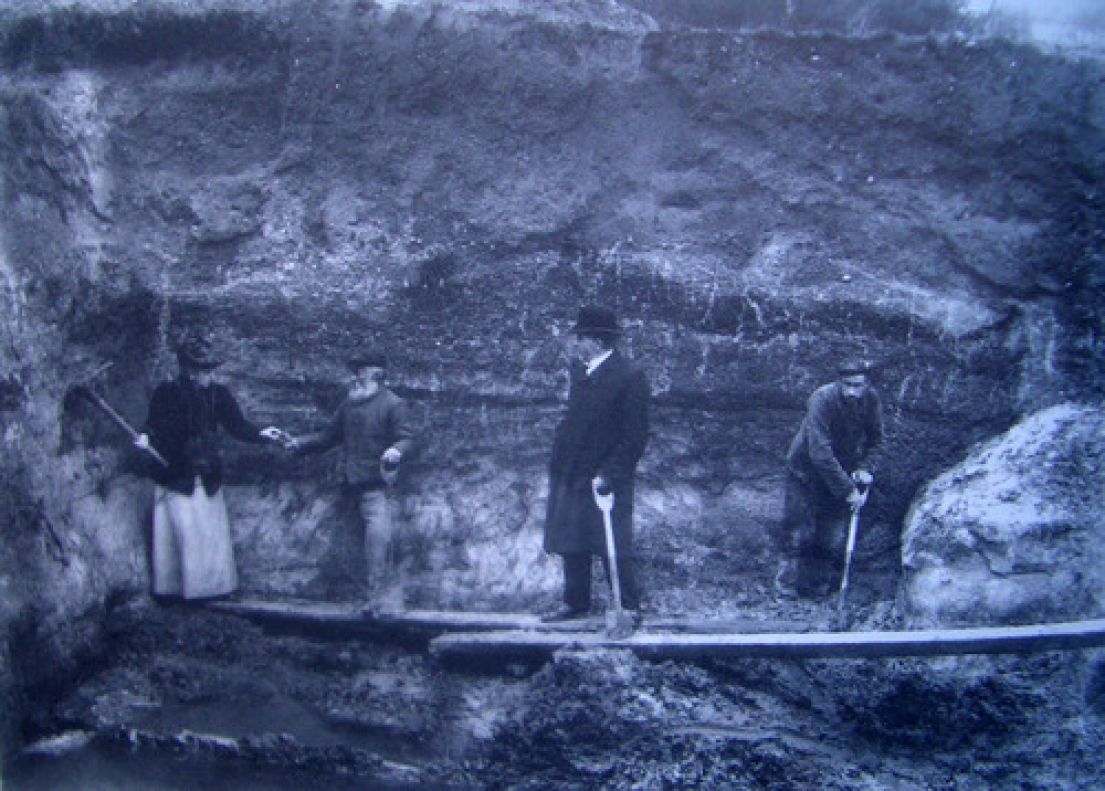 A black and white photograph of men in bowler hats holding shovels as well as a women in a Victorian style dress in an archaeological pit/